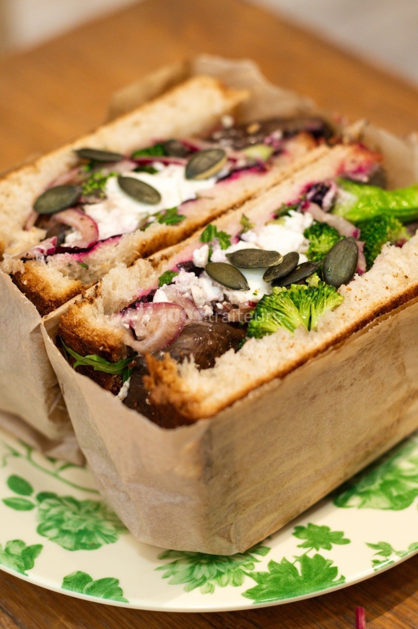 Sandwich végétarien garni de légumes, fromage frais, graines de courge et herbes, présenté dans un emballage papier.