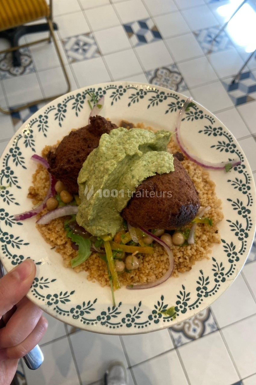 Assiette de couscous garnie de falafels, légumes, pois chiches et sauce verte, servie sur une table carrelée.