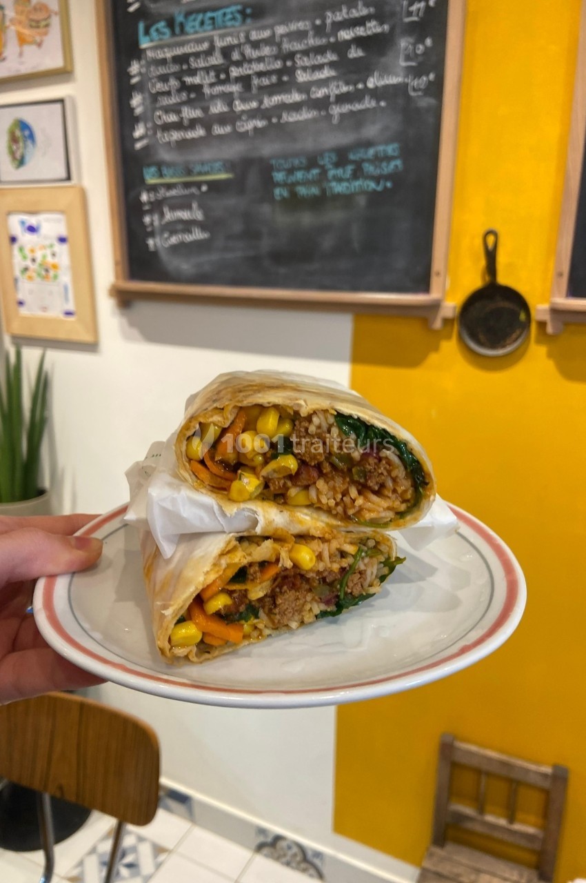 Un burrito végétarien garni de légumes, maïs et quinoa, présenté coupé en deux sur une assiette dans un café coloré.