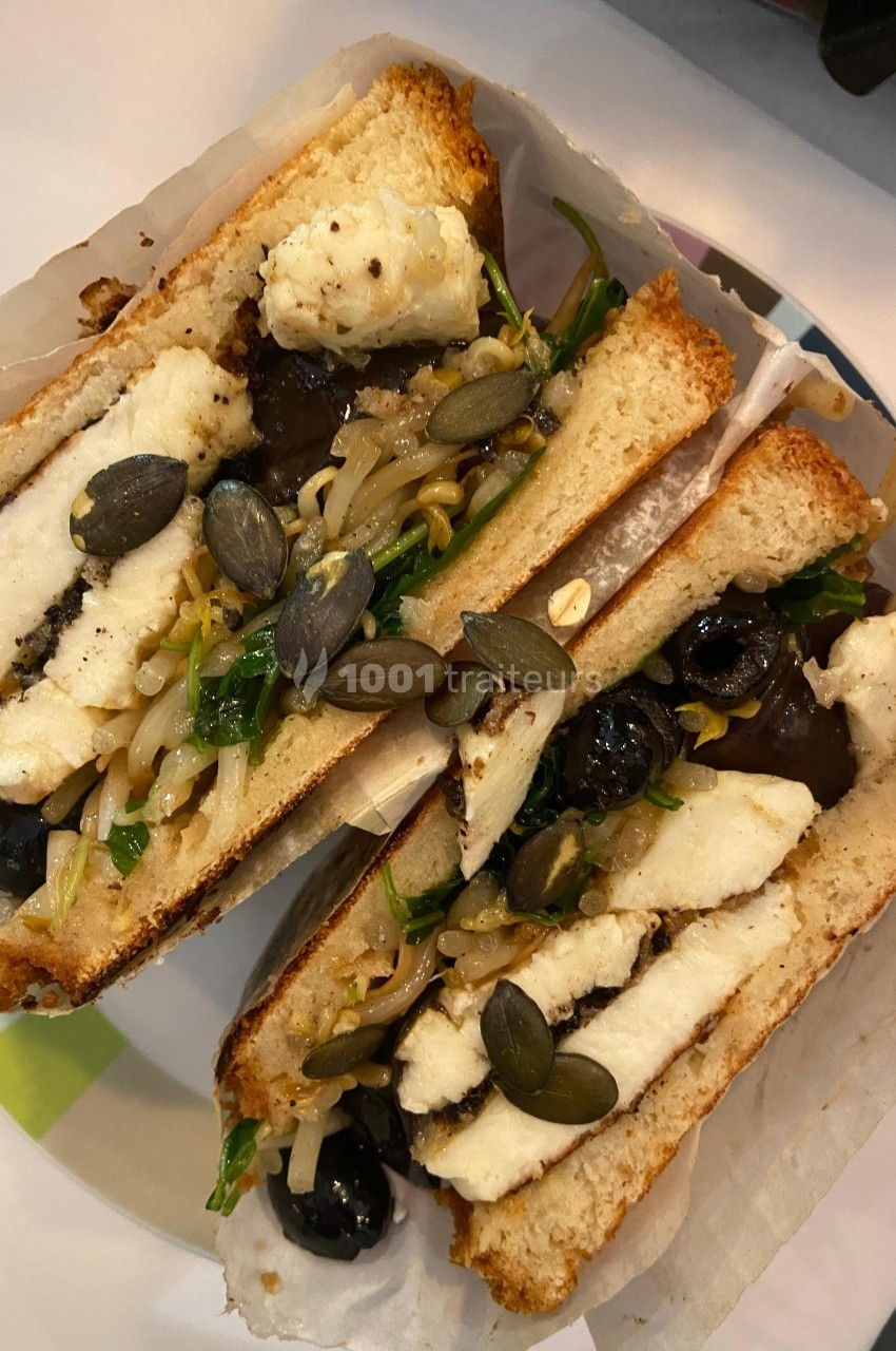 Sandwich garni de fromage grillé, légumes sautés, graines de courge et olives noires, sur pain toasté.