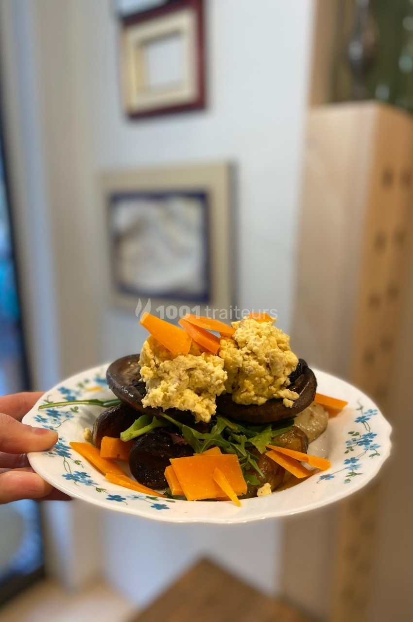 Assiette garnie de légumes grillés, roquette, morceaux de fromage et une préparation crémeuse, tenue à la main.