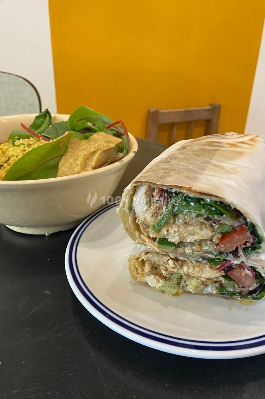 Un wrap garni de légumes et sauce, accompagné d'un bol de salade et houmous, posé sur une table noire.