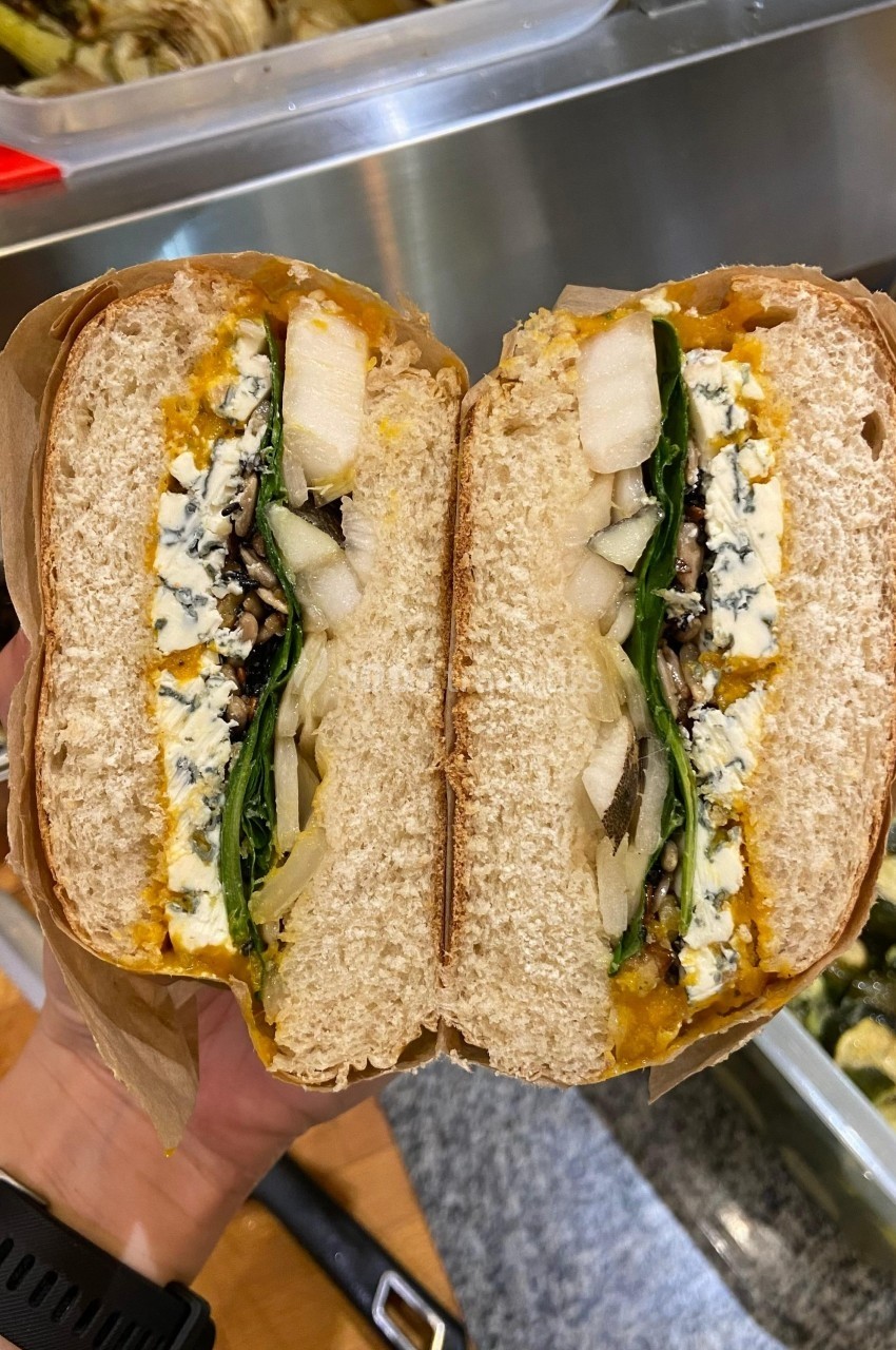 Sandwich coupé en deux, garni de fromage bleu, poires, épinards et autres ingrédients, tenu au-dessus d'un plan de travail.