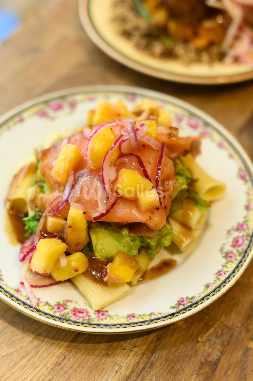 Assiette de pâtes garnie de saumon fumé, avocat, ananas, oignons rouges et sauce sur une table en bois.