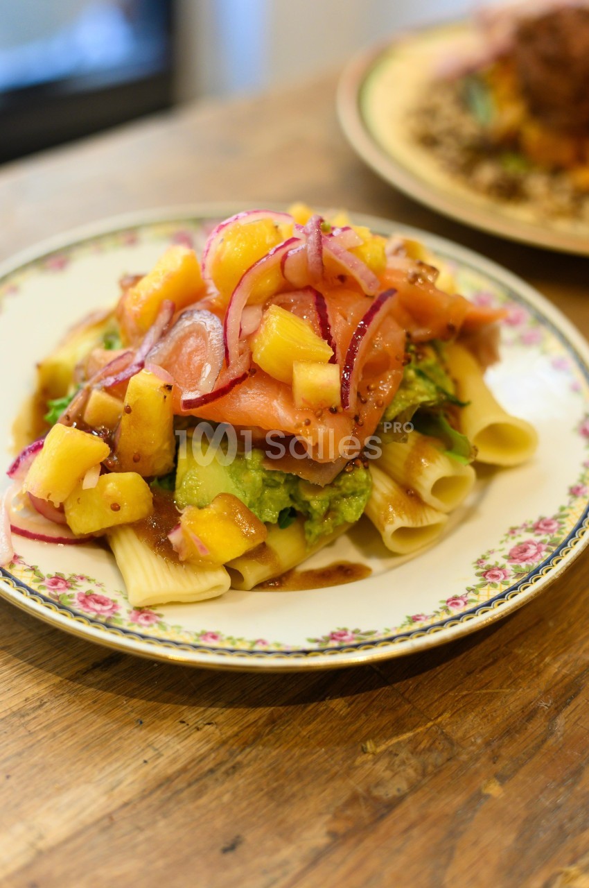 Assiette de pâtes garnie de saumon fumé, avocat, morceaux de mangue et oignons rouges sur une table en bois.