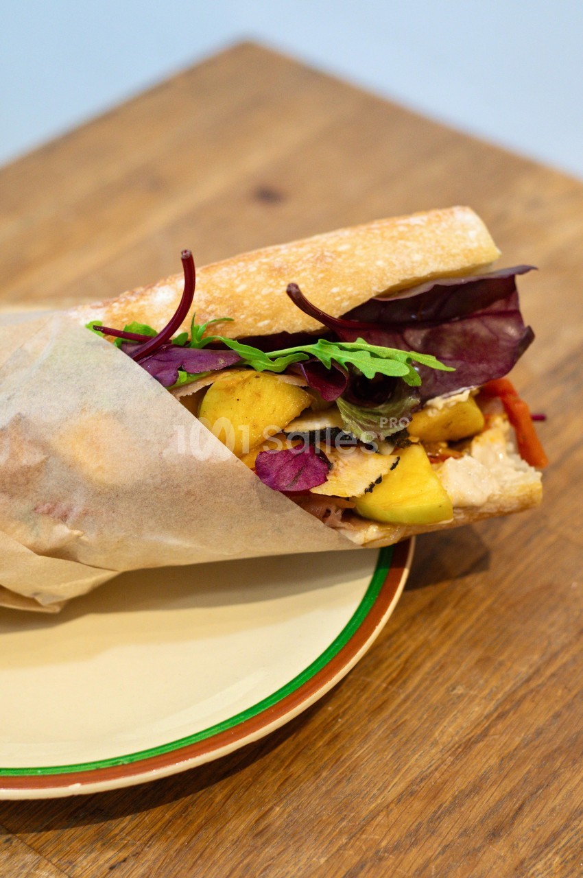 Sandwich garni de poulet, feuilles de salade, morceaux de mangue et légumes, présenté sur une assiette en bois.