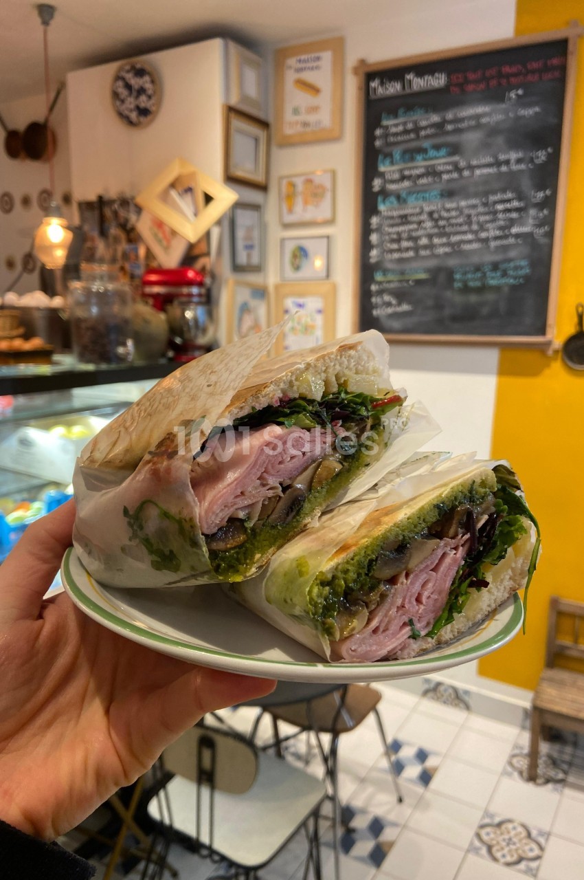 Un sandwich garni de jambon, légumes et sauce pesto, présenté coupé en deux dans un café chaleureux.