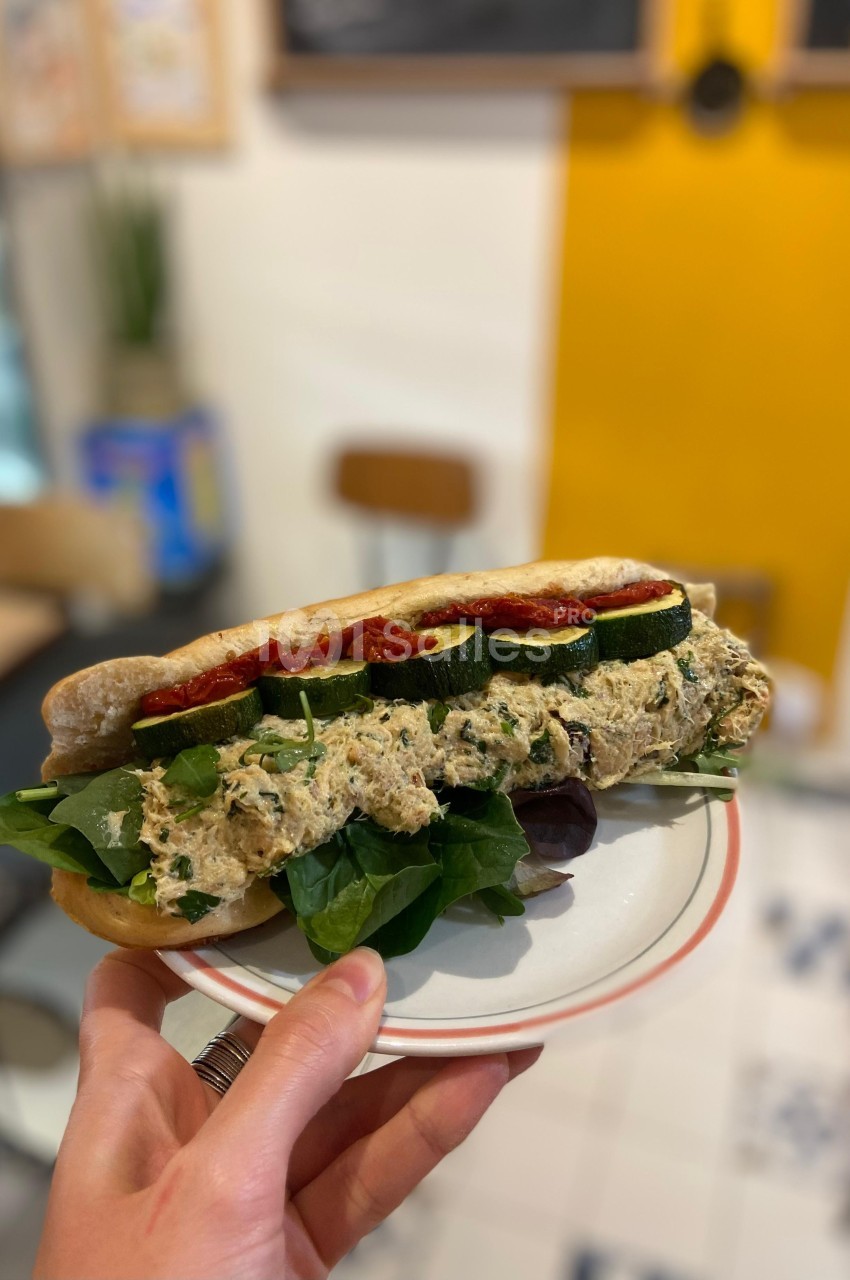 Sandwich garni de légumes grillés, feuilles de salade et tartinade, tenu sur une assiette dans un café lumineux.