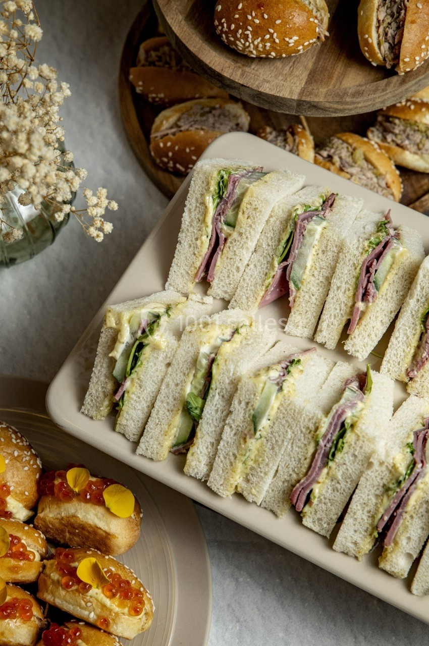 Plateau de sandwichs garnis de légumes et charcuterie, accompagné de petits pains et amuse-bouches sur une table.