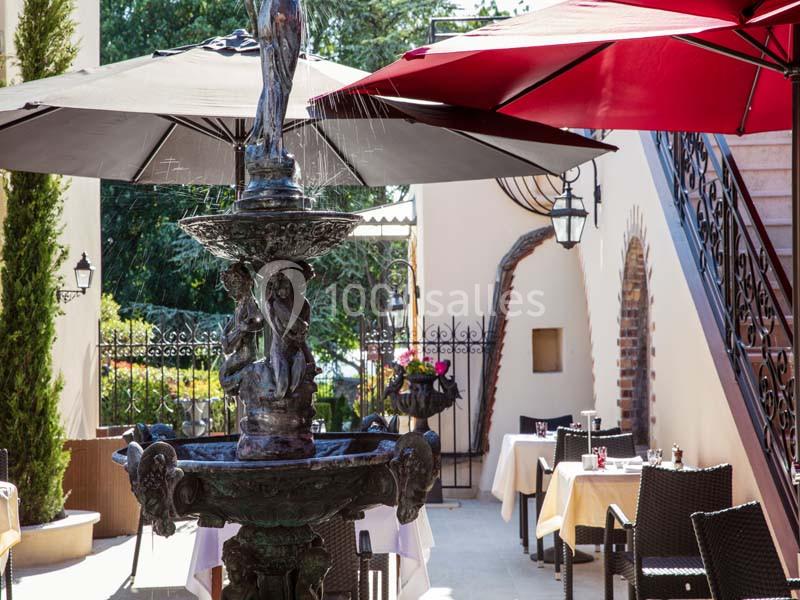 Fontaine décorative en métal entourée de tables de restaurant en extérieur, avec parasols rouges et noirs.