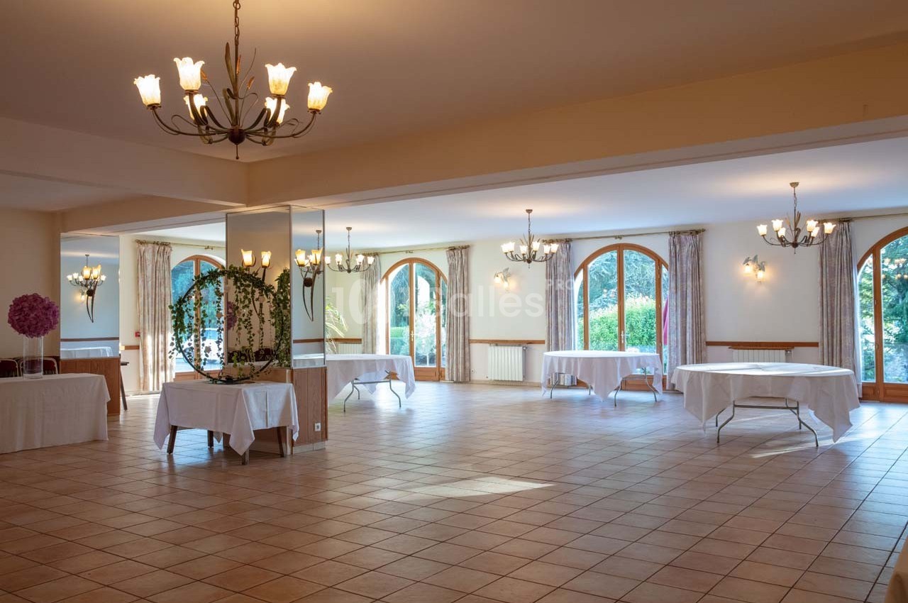 Salle lumineuse avec grandes fenêtres cintrées, tables rondes nappées blanches et lustres suspendus.