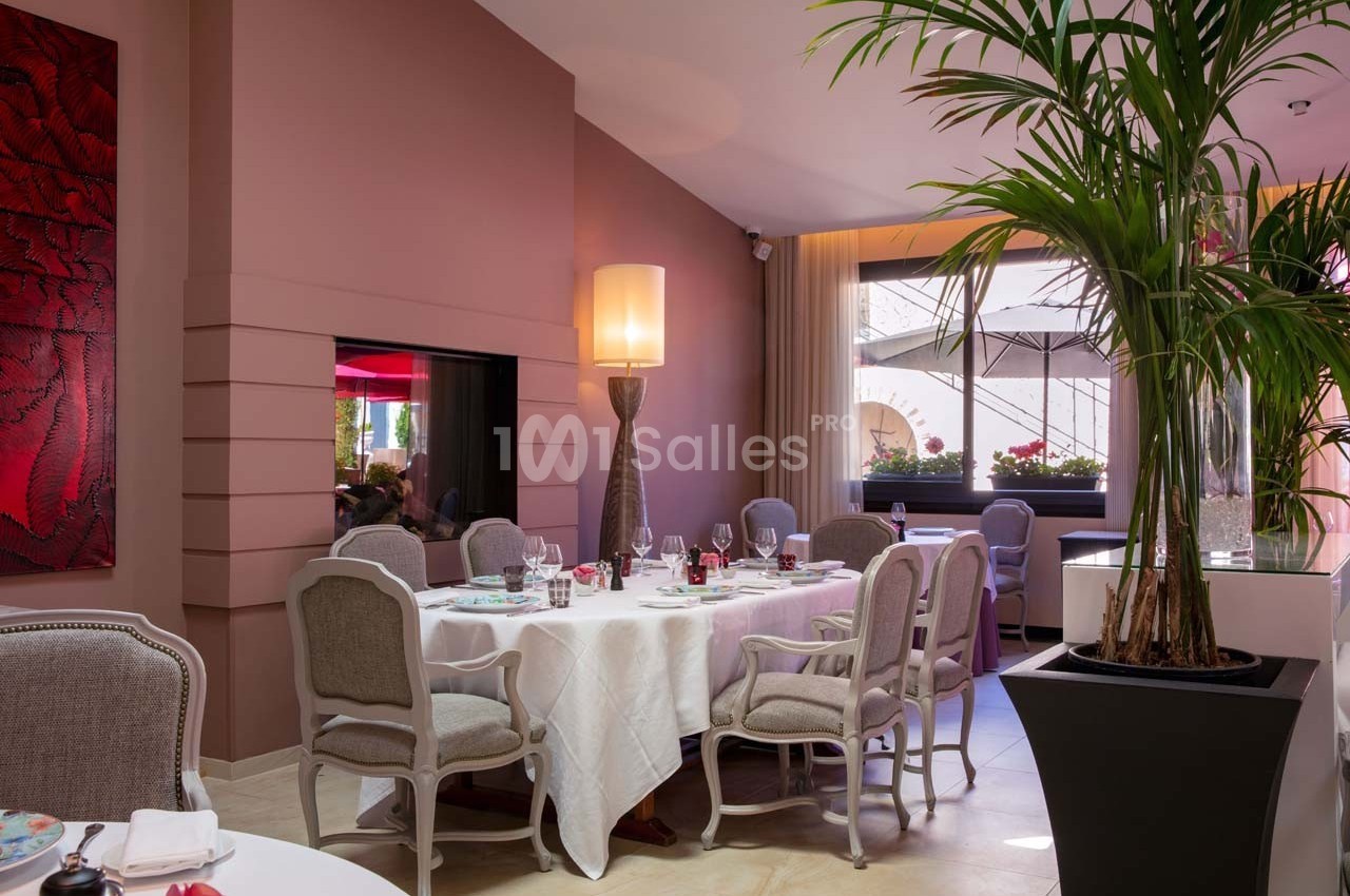 Salle de restaurant élégante avec tables dressées, chaises rembourrées, grande plante et lumière naturelle.