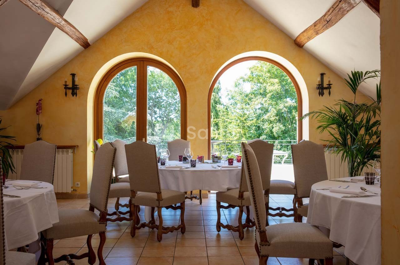 Salle lumineuse avec tables dressées, chaises en bois et grandes fenêtres en arche donnant sur un jardin verdoyant.