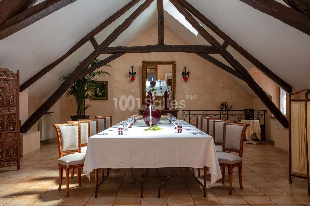 Salle à manger sous des poutres apparentes avec une grande table dressée, entourée de chaises élégantes.