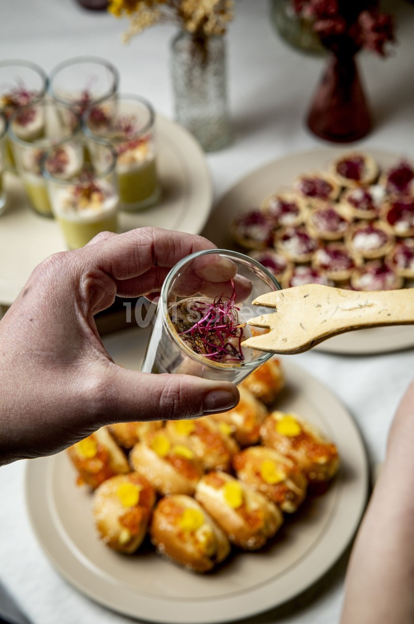 Une main tient un verre de verrine décorée, entourée d'assiettes de bouchées et d'amuse-bouches variés.