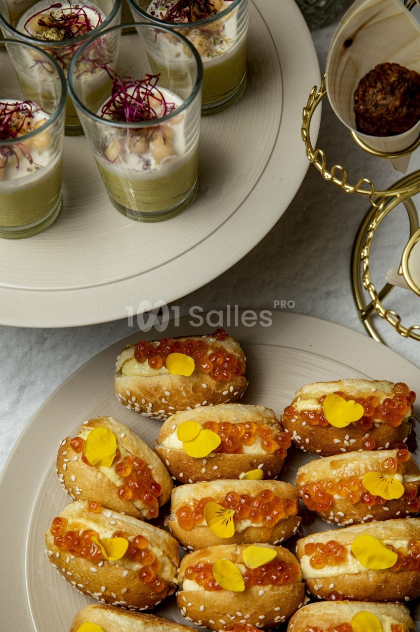 Plateau de mini hot-dogs garnis d'œufs de poisson et fleurs comestibles, avec verrines en arrière-plan.