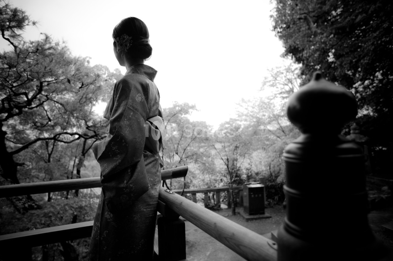 Femme en kimono vue de dos, debout sur une terrasse en bois, entourée d'arbres dans un paysage naturel.