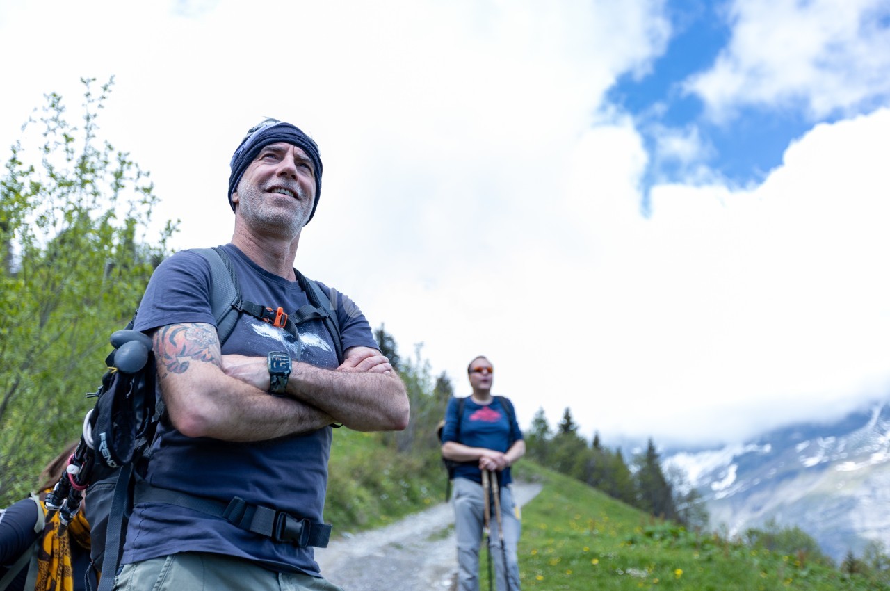 Un homme souriant avec un sac à dos sur un sentier de montagne, accompagné d'une autre personne en arrière-plan.