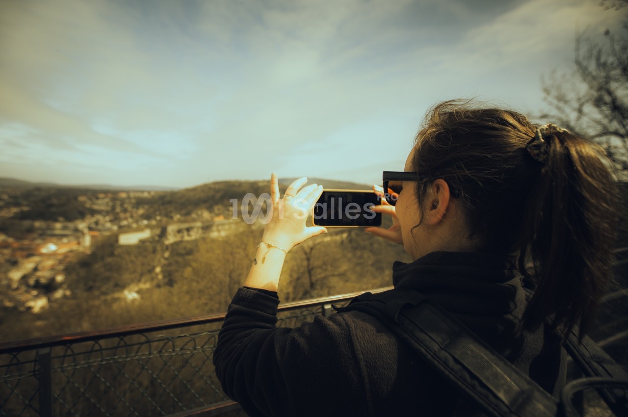 Une femme photographie un paysage vallonné depuis un point de vue, appuyée contre une barrière métallique.