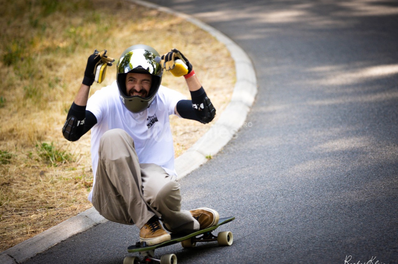 Un skateur en descente sur une route, portant un casque et des protections, fait un geste amusant avec les mains.