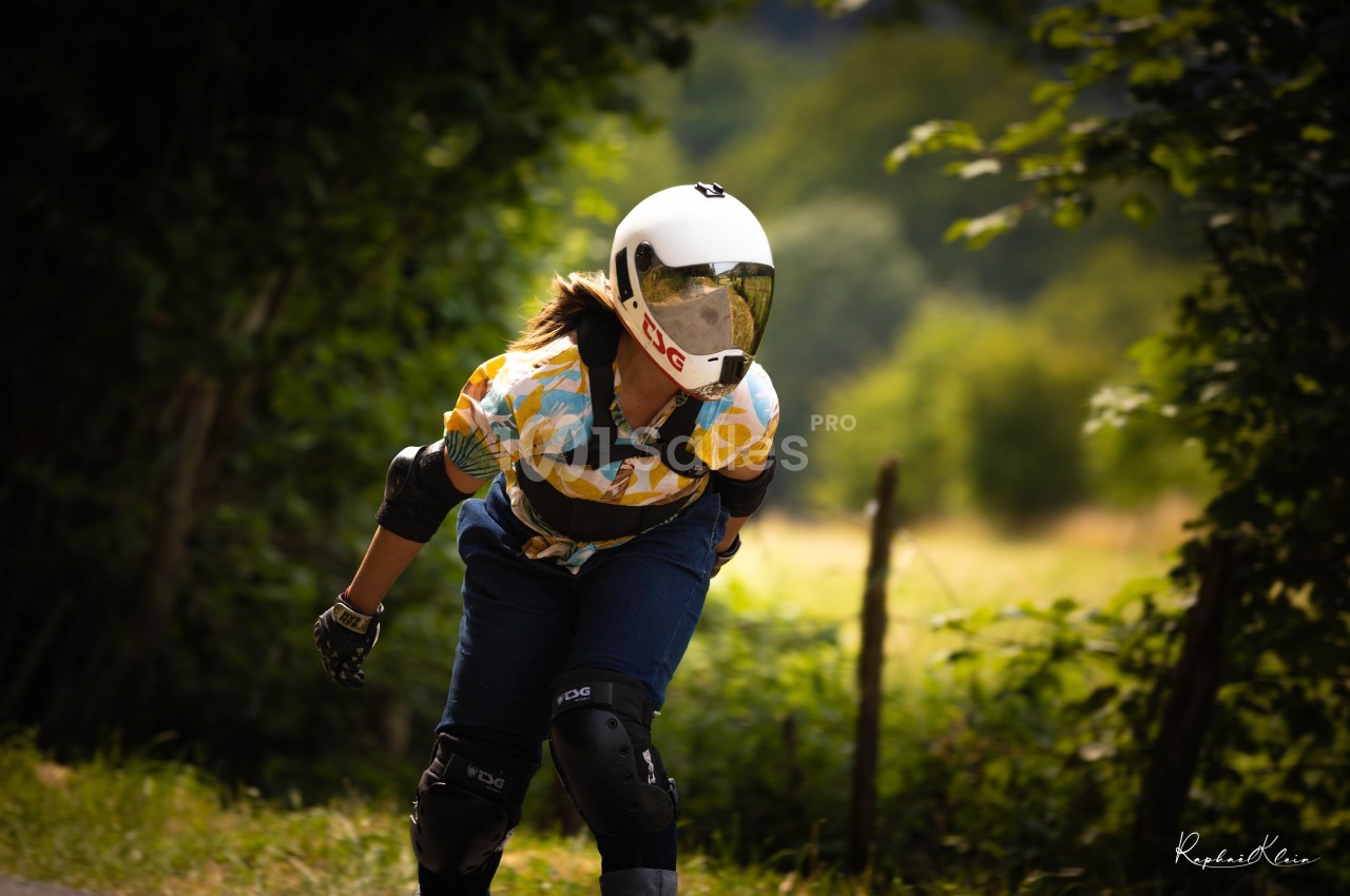 Personne portant un casque et des protections, pratiquant le longboard sur une route bordée de verdure.