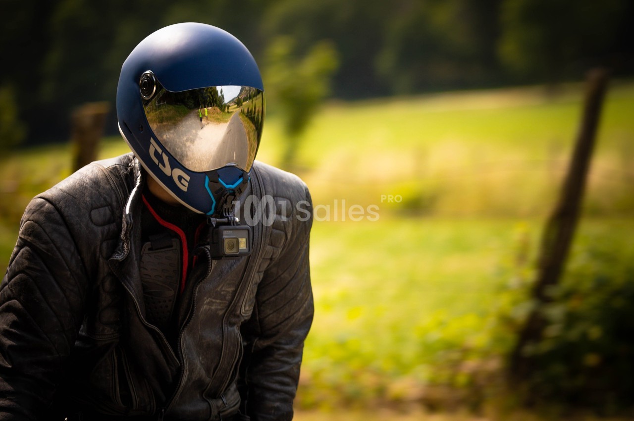 Motard portant un casque bleu avec visière réfléchissante, équipé d'une caméra, dans un paysage champêtre flou.