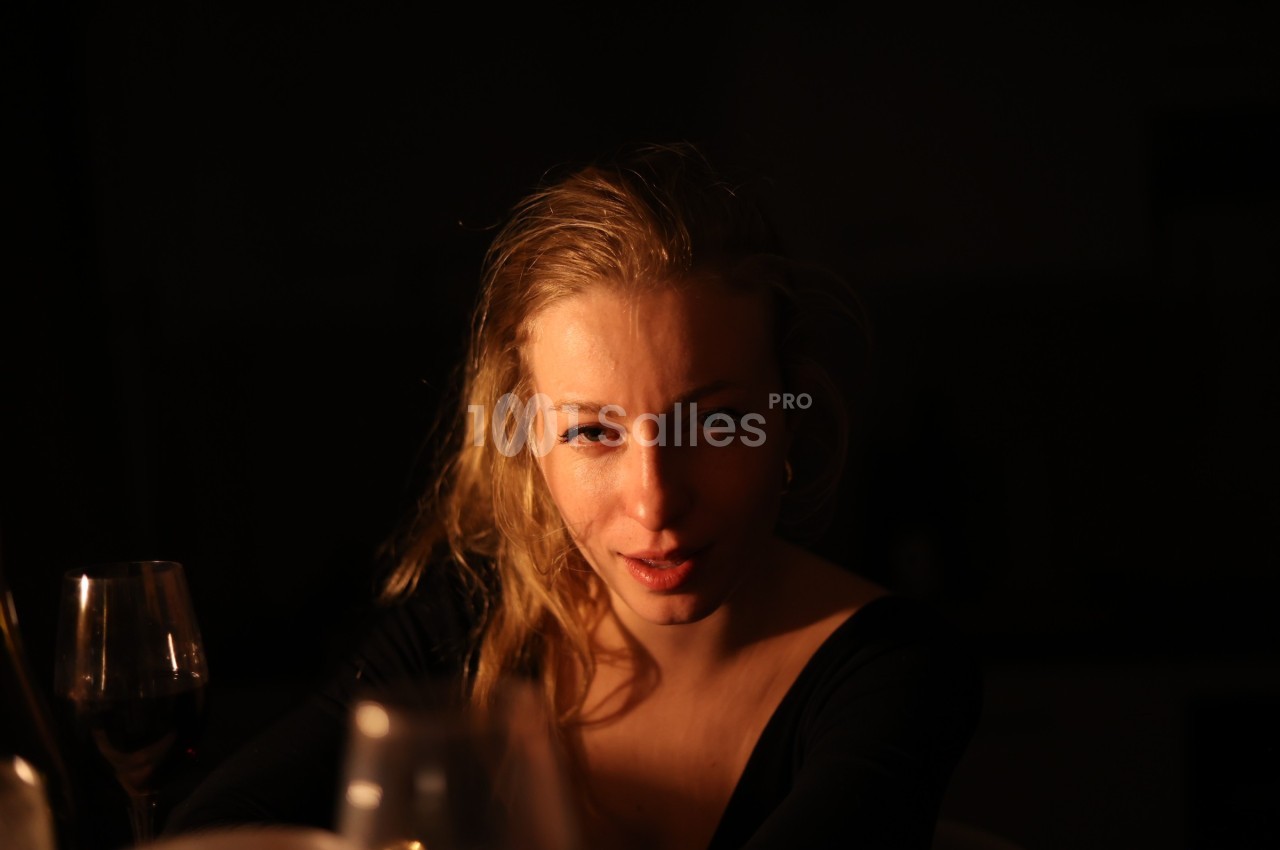Femme assise dans une lumière tamisée, entourée de verres, regardant vers l'objectif.