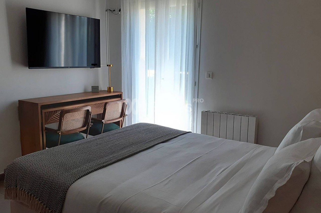 Chambre lumineuse avec lit double, télévision murale, bureau en bois, deux chaises et rideaux blancs filtrant la lumière.
