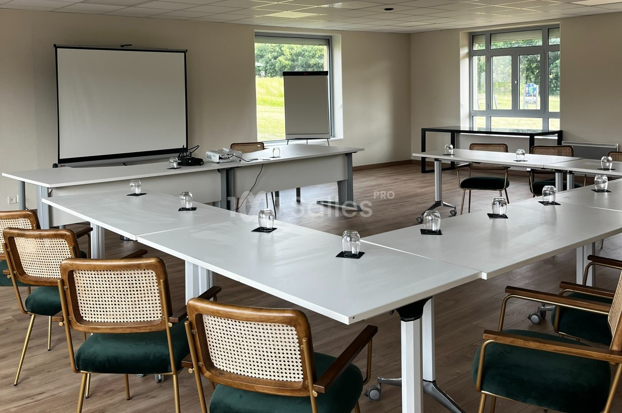 Salle de réunion lumineuse avec tables disposées en U, chaises en bois et écran de projection à l'avant.