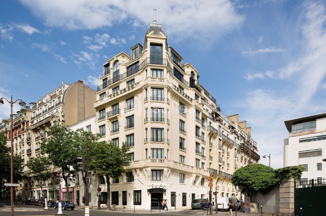 Immeuble haussmannien d'angle avec balcons, situé dans une rue urbaine bordée d'arbres sous un ciel dégagé.