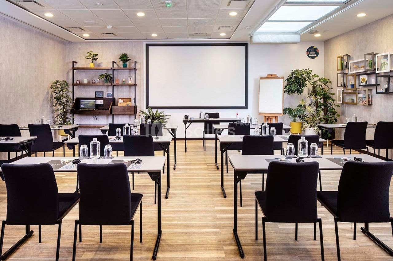 Salle de réunion lumineuse avec des tables et chaises alignées, un écran de projection et des plantes décoratives.