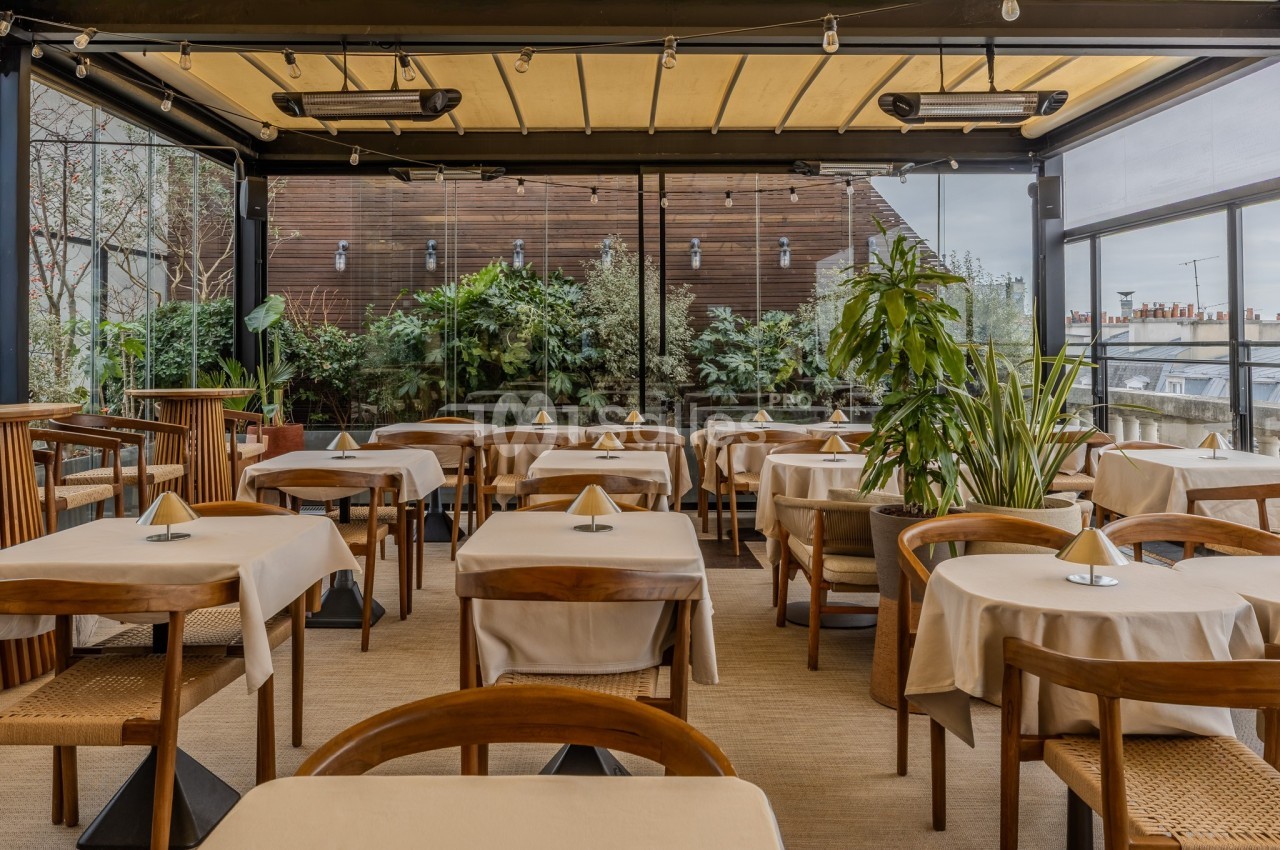 Salle de restaurant lumineuse avec tables dressées, chaises en bois et grandes baies vitrées donnant sur des plantes.