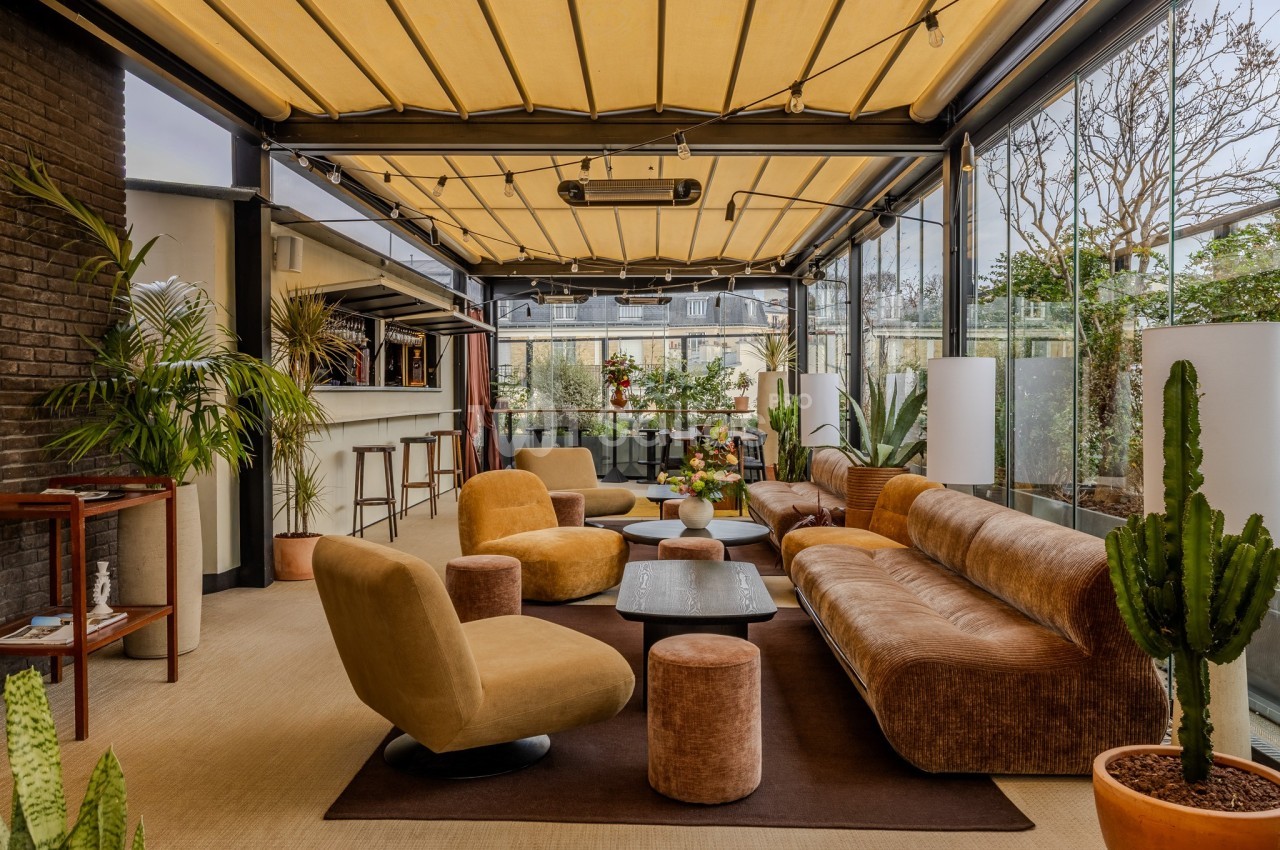 Espace lounge lumineux avec canapés, fauteuils, tables basses et plantes, sous une verrière avec toit ouvrant.