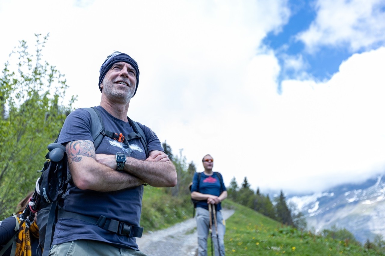 Un homme souriant en tenue de randonnée, bras croisés, sur un sentier de montagne par temps ensoleillé.