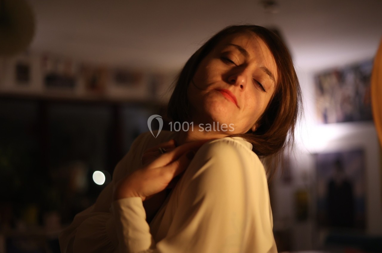 Femme aux yeux fermés dans une pièce faiblement éclairée, portant une blouse claire et semblant pensive.