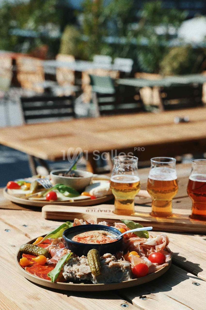 Plateau de charcuterie et légumes avec une sauce au centre, accompagné de bières sur une table en terrasse ensoleillée.