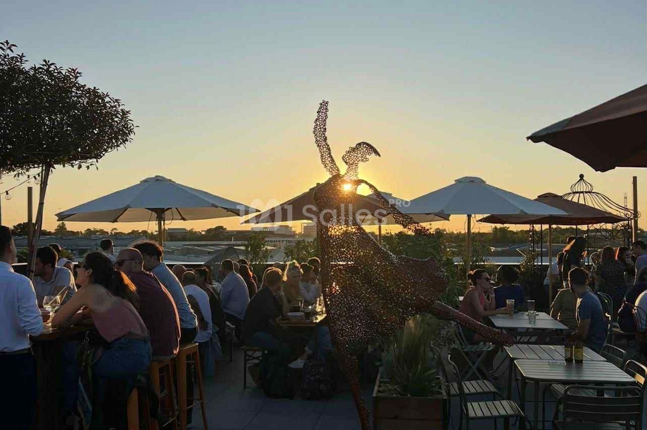 Statue d'une danseuse en métal sur une terrasse animée au coucher du soleil, entourée de parasols et de convives.