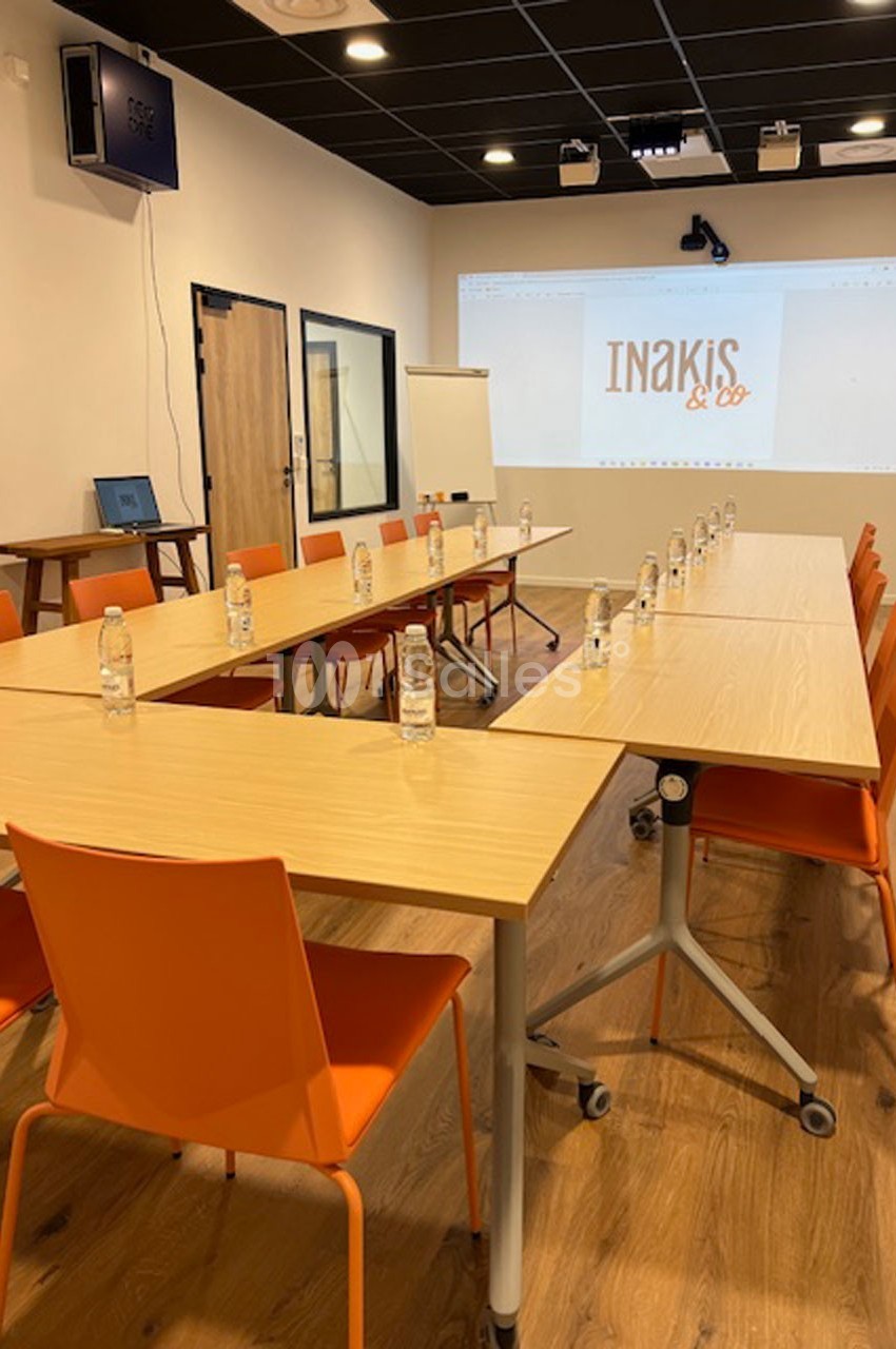 Salle de réunion équipée de tables en bois clair, chaises orange, bouteilles d'eau et écran de projection allumé.