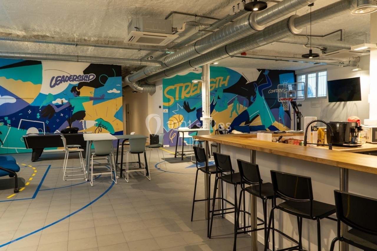 Espace intérieur moderne avec tables, chaises hautes, muraux colorés et panier de basketball en arrière-plan.