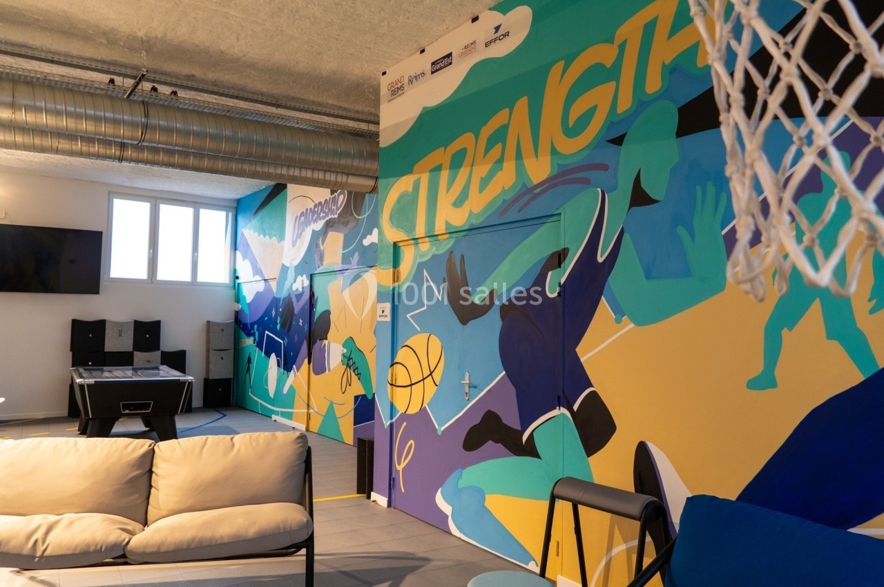 Mur coloré avec fresque sportive représentant des joueurs de basket, dans une salle équipée de canapés et d'une table de jeu.