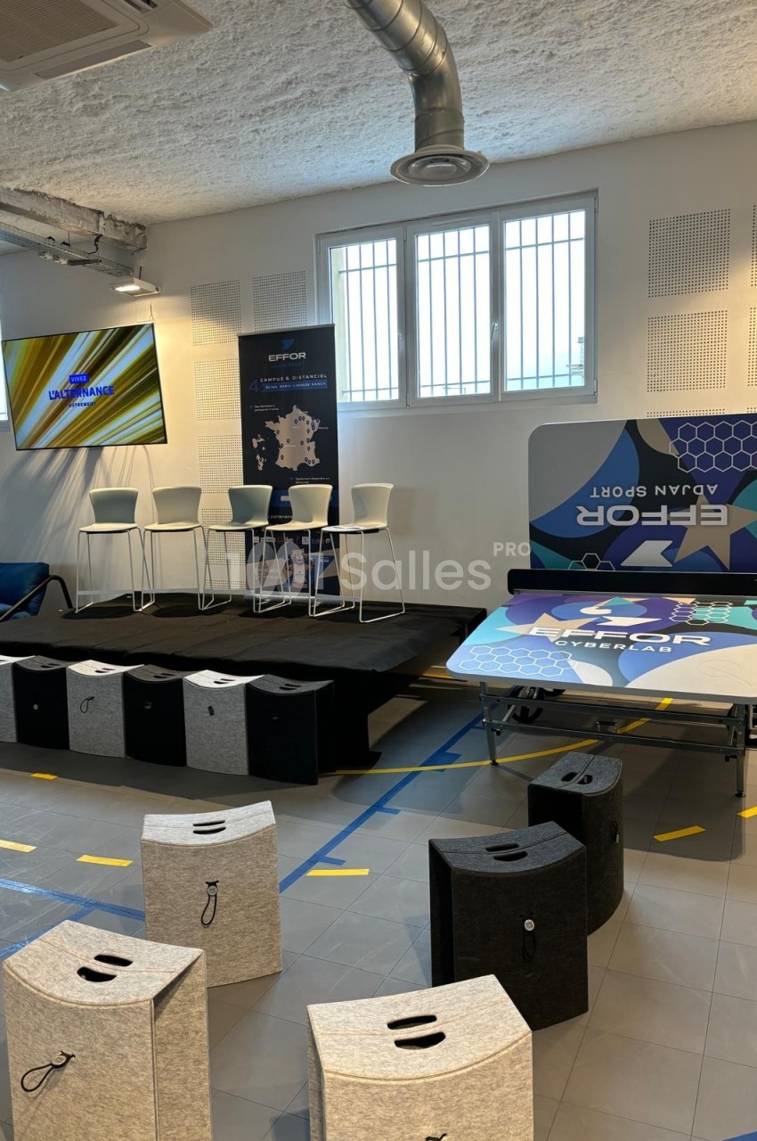 Salle de conférence moderne avec estrade, chaises hautes, affiches promotionnelles et tabourets en premier plan.