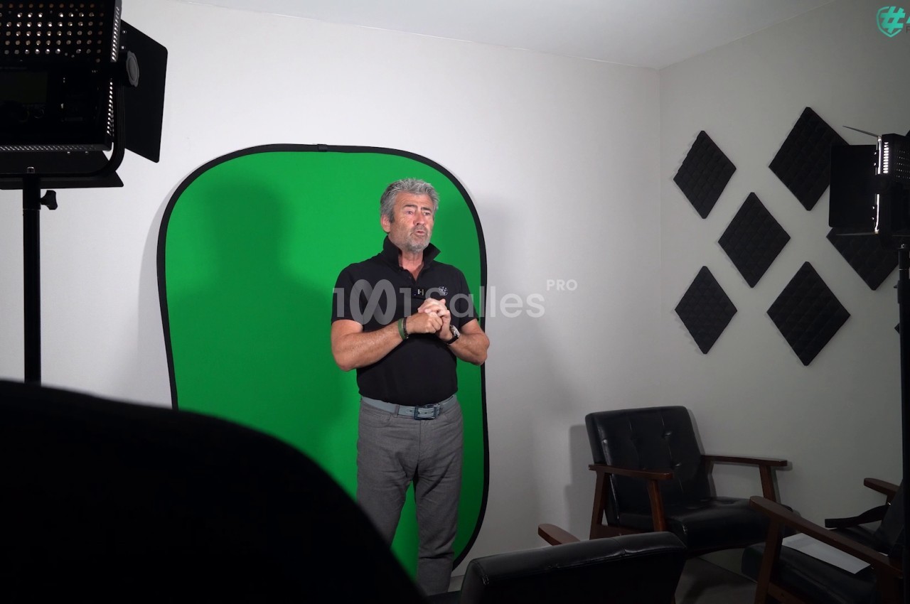 Un homme debout dans un studio avec un fond vert, éclairé par des projecteurs, entouré de panneaux acoustiques.