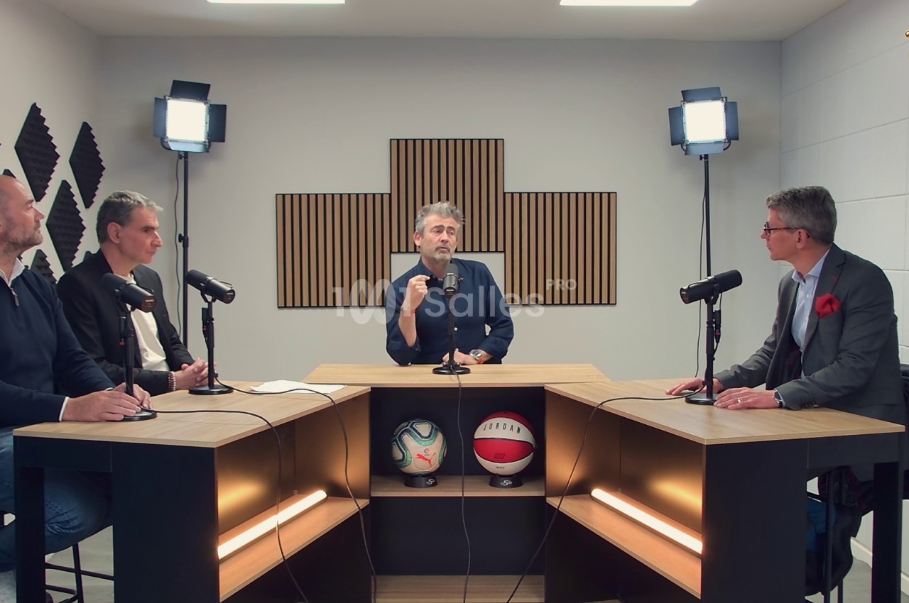 Quatre hommes discutent autour de tables équipées de micros dans un studio avec éclairage et panneaux acoustiques.