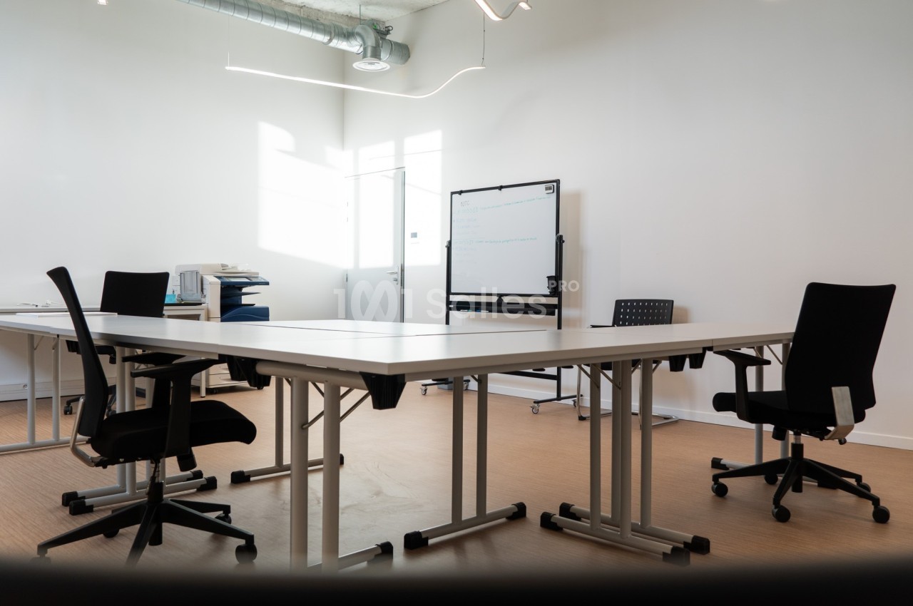 Salle de réunion lumineuse avec tables blanches, chaises noires, tableau blanc et équipement de bureau.