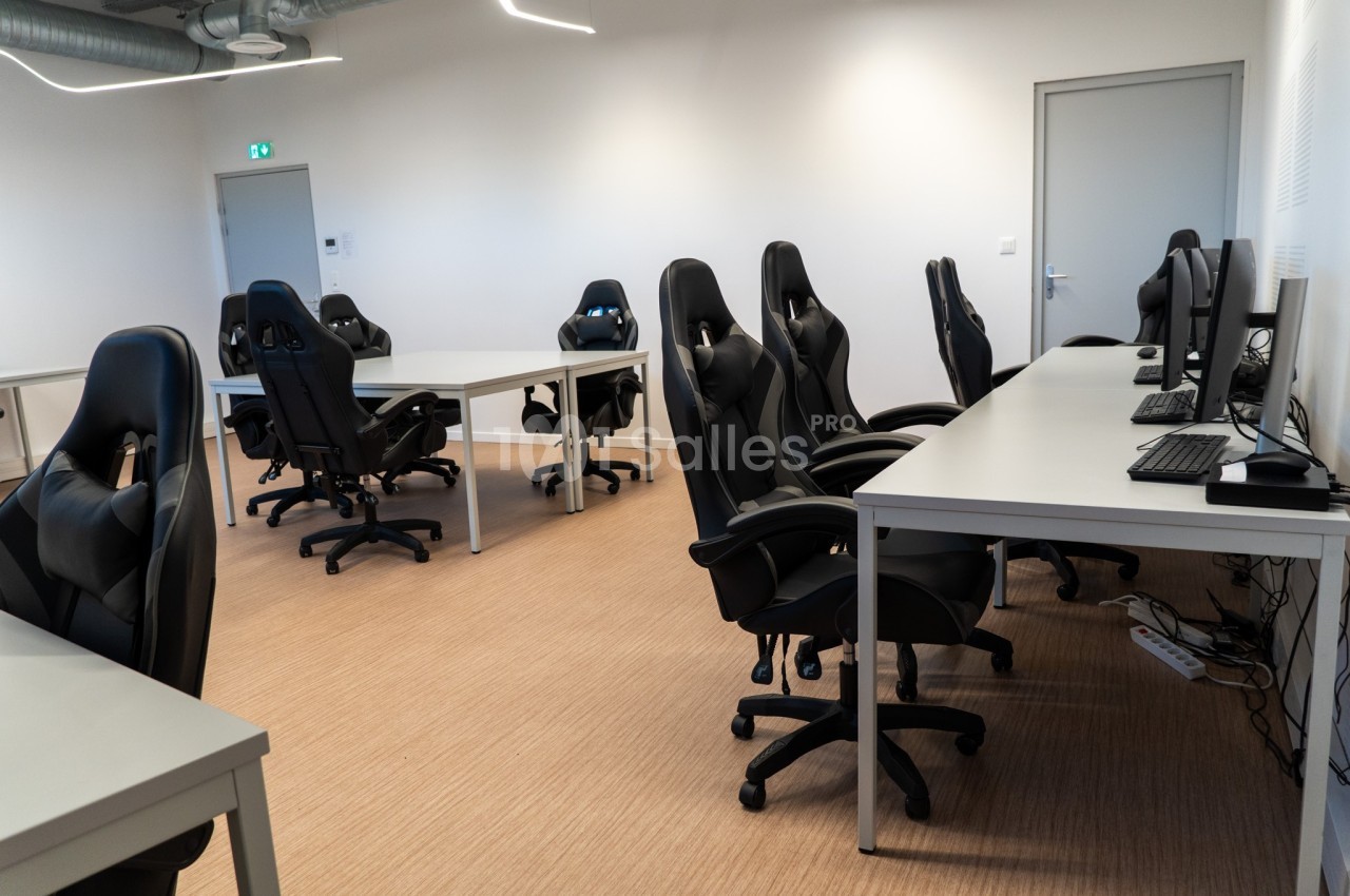 Salle équipée de bureaux, chaises ergonomiques et ordinateurs, éclairée par des néons, avec un sol en parquet clair.