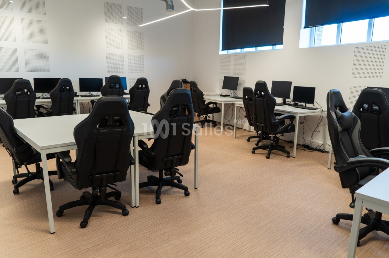 Salle équipée de bureaux avec ordinateurs et chaises ergonomiques noires, éclairée par des fenêtres partiellement occultées.