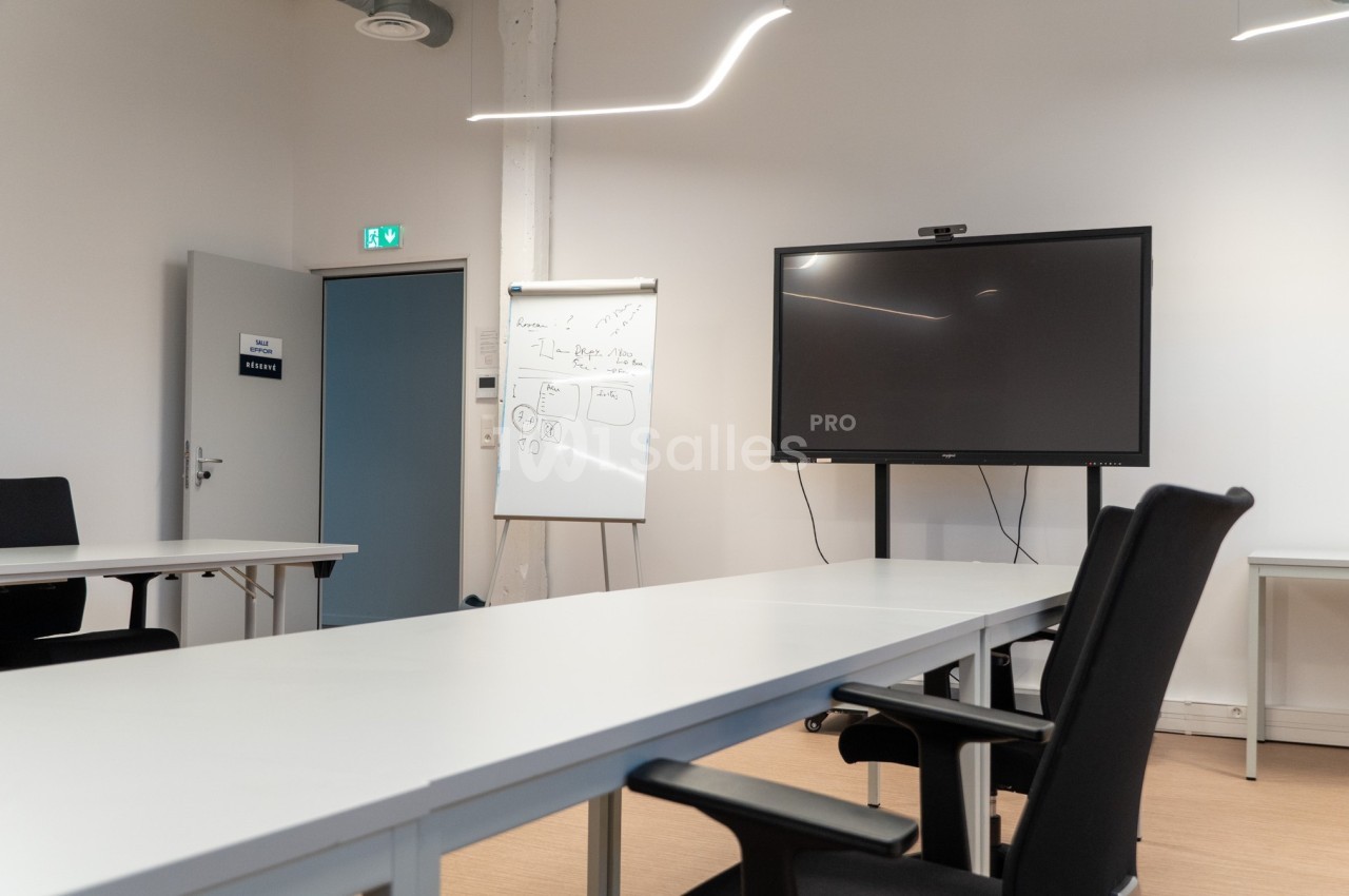 Salle de réunion moderne avec table blanche, chaises noires, tableau blanc et écran de présentation.