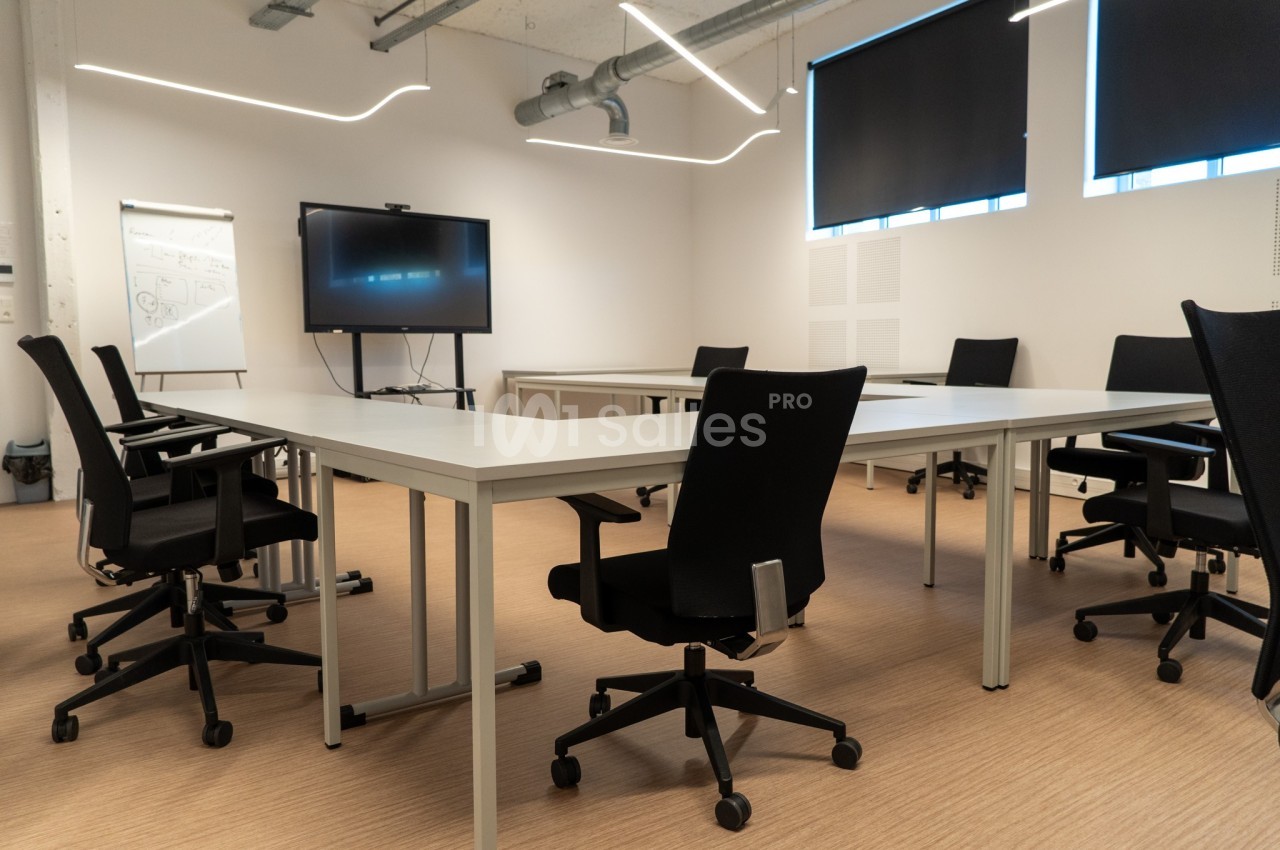 Salle de réunion moderne avec des tables blanches, des chaises noires, un tableau blanc et un écran de présentation.