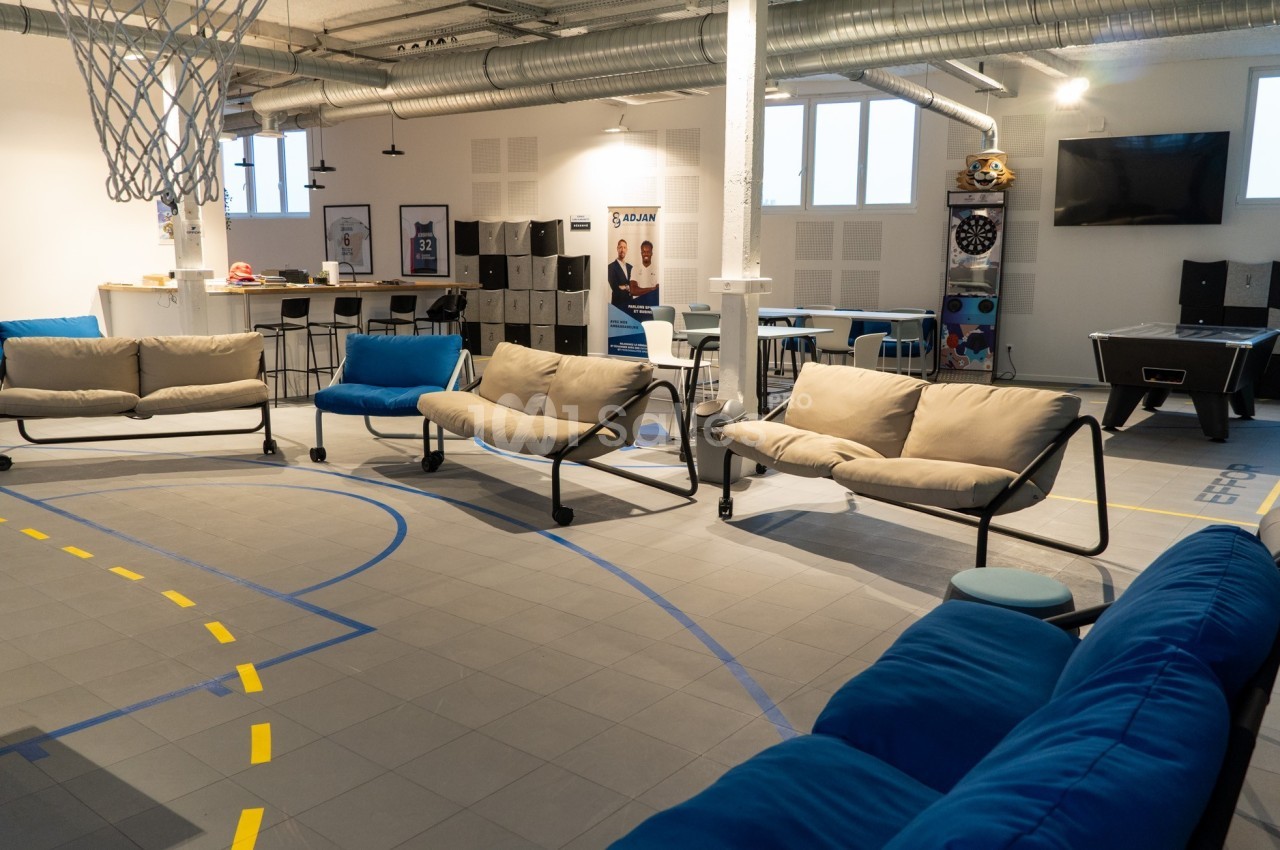 Salle de détente avec canapés, baby-foot, fléchettes, panier de basket et coin cuisine dans un espace lumineux.