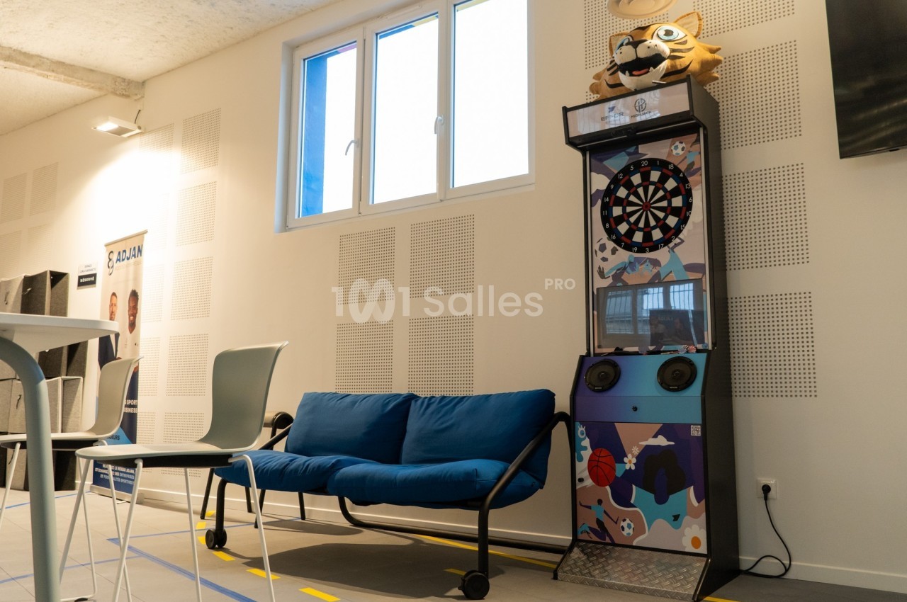 Un canapé bleu et une cible de fléchettes décorée dans une salle lumineuse avec des murs blancs.